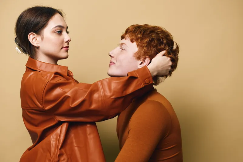 Woman and man in brown outfits looking at each other with affection on beige background. Couple embracing in warm clothes with casual style and intimate connection in studio light.