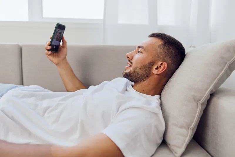 Relaxed man using smartphone at home, engaging in a video call. Bright and modern living room setting reflects a cozy lifestyle and technology integration.