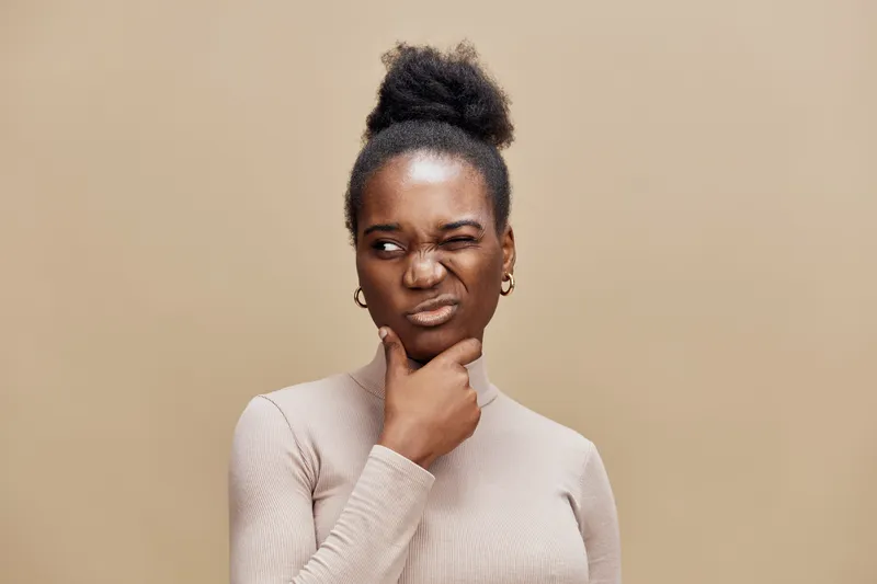 Thoughtful Black woman pondering a question with a skeptical expression on a neutral beige background, dressed in a light turtleneck sweater