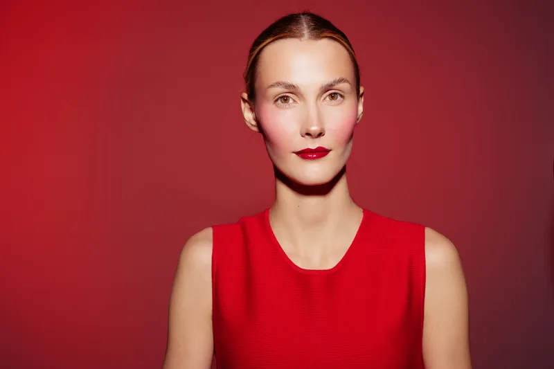Portrait of confident woman with red lipstick and red dress on red background. Studio lighting highlights her clear skin and calm expression. Concept of beauty and fashion.