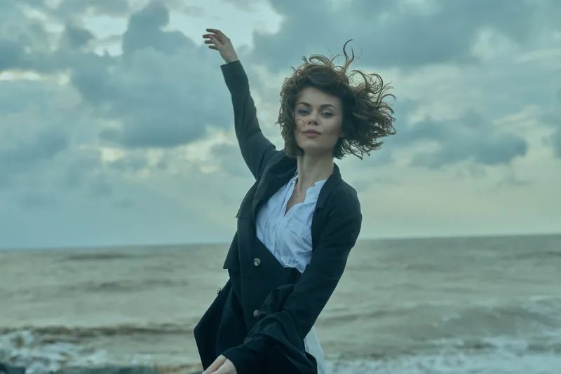 Woman in dark blazer and white shirt dances by the shoreline, hair and jacket fluttering as ocean waves roll beneath a cloudy sky, creating a dynamic coastal mood and confident style.