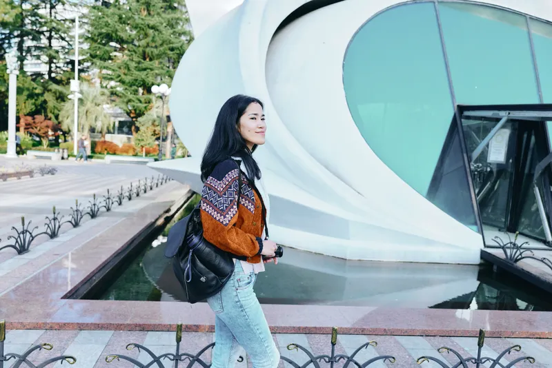 Woman walking outdoors beside futuristic architecture, stylish orange patterned top and jeans, carrying a backpack, urban scenery, daytime light, calm mood, travel vibe, candid street shot