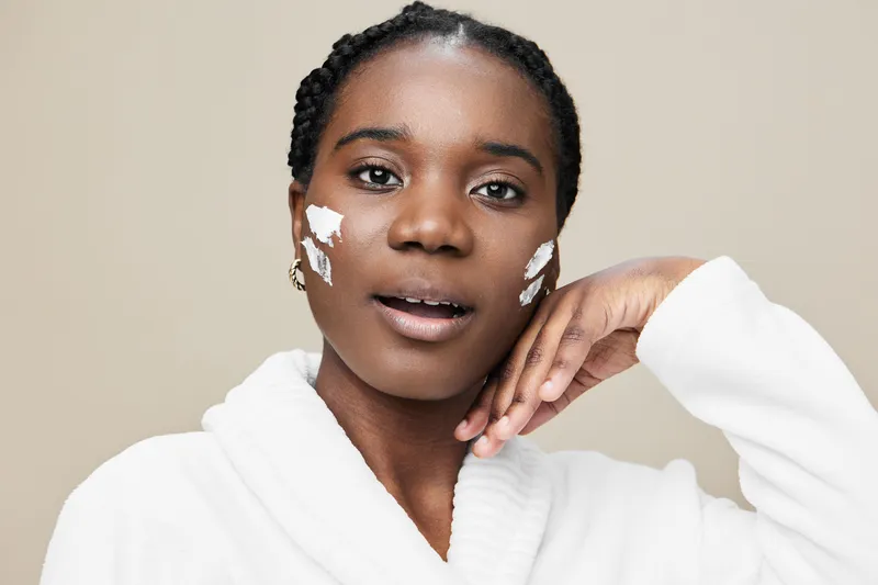 Stylish young Black woman with skincare cream on her face, wearing a white robe, presenting a serene expression against a neutral beige background