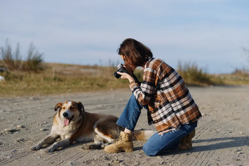 A photographer kneels on a sandy beach, capturing a playful dog with a camera, wrapped in a plaid shawl and denim, under wide sky and peaceful coastal scenery.