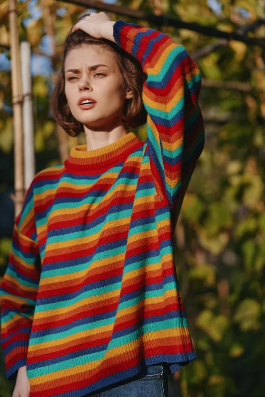 Young adult woman in rainbow sweater with short hair, squinting and leaning with hand on head in sunlit outdoor garden. Casual outfit, autumn lifestyle portrait for fashion and travel editorial.