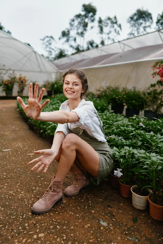 Young woman with dirty hands and a joyful smile, sitting in a greenhouse surrounded by vibrant green plants and flowers, showcasing a love for gardening