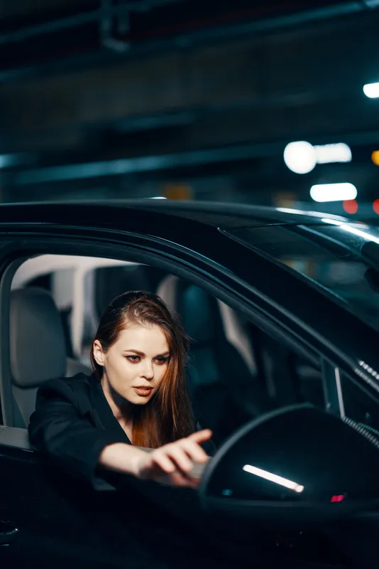 Вертикальный снимок сбоку, ночь: женщина сидит в автомобиле, смотрит в окно и тянется рукой к боковому зеркалу заднего вида