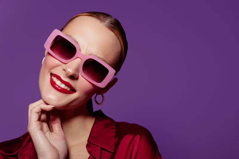 Smiling woman with pink sunglasses and red lipstick poses against purple background. Fashion portrait with stylish accessories and confident expression in studio lighting.