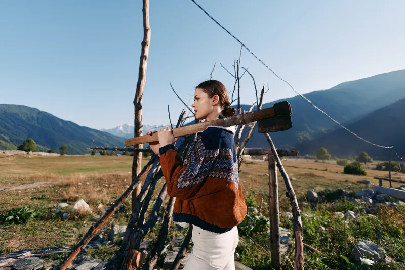 Woman carrying a wooden log on her shoulder in countryside field by a rustic fence, wearing a patterned sweater with mountains in the background, rural outdoor portrait.