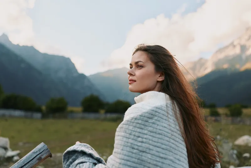Woman wrapped in a soft blanket outdoors, with distant mountains and bright sky behind. Concept of relaxation, nature, and wellness. Soft lighting highlights features and mood.