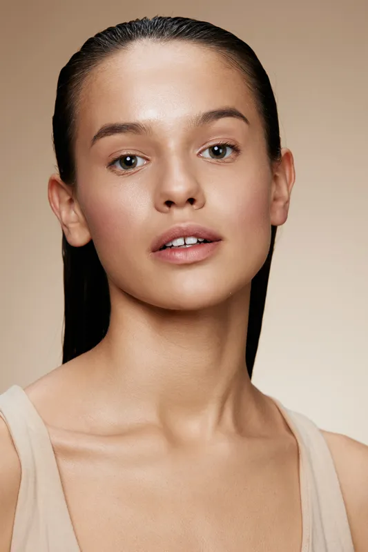 Natural beauty portrait of a young woman with wet hair and minimal makeup, showcasing healthy skin and a neutral background that enhances her features