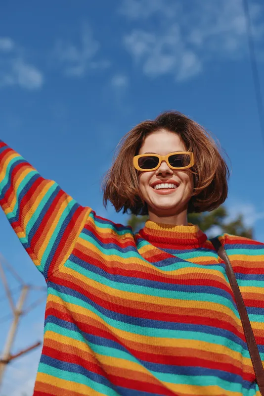 Woman smiling in rainbow sweater and orange sunglasses, short brown hair, mid-20s, arms raised outdoors under blue sky, joyful casual fashion portrait for lifestyle and travel.