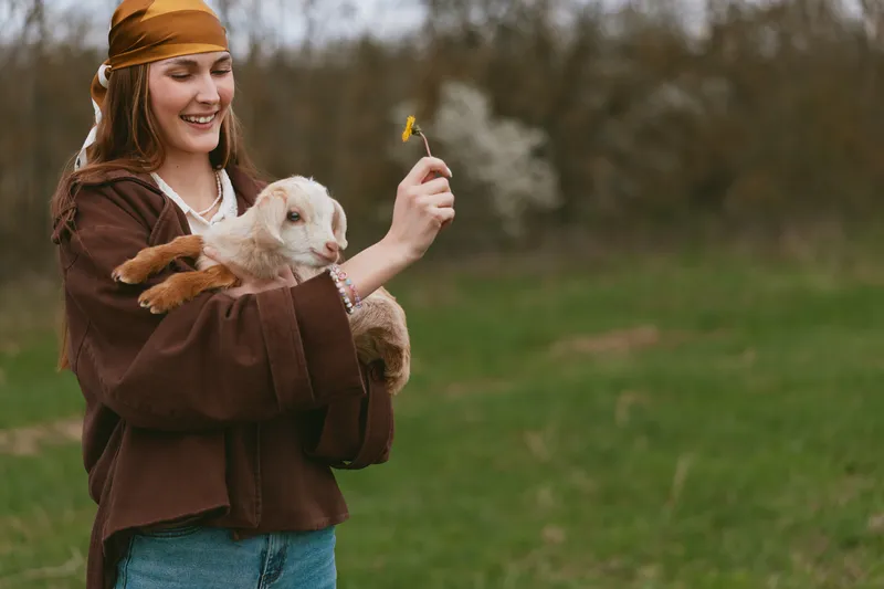 Smiling woman in rustic clothing holds a goat kid outdoors, offering a flower. Soft natural light on a grassy meadow expresses care, companionship, wellness, and lifestyle moments.