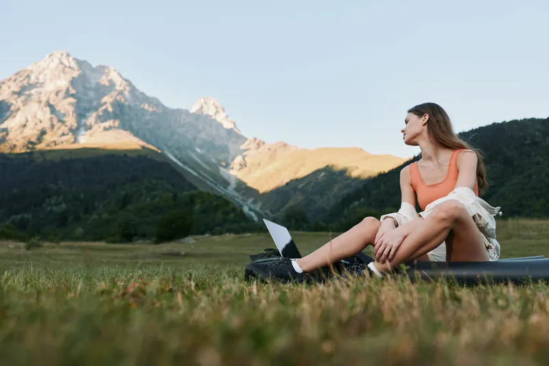 An outdoor scene shows a woman seated on grass with a laptop, gazing into the distance. She looks focused yet relaxed, creating a calm, productive mood in a natural setting.