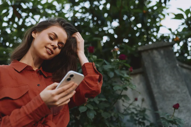 A smiling woman in a rust red shirt uses a smartphone outdoors among garden foliage. A casual lifestyle portrait capturing nature, leisure, and everyday technology