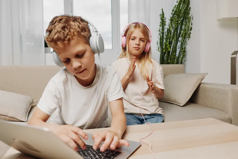 Two children at home, boy focused on laptop and girl enjoying music with headphones, casual clothing in bright living room. Concept of digital play and leisure.