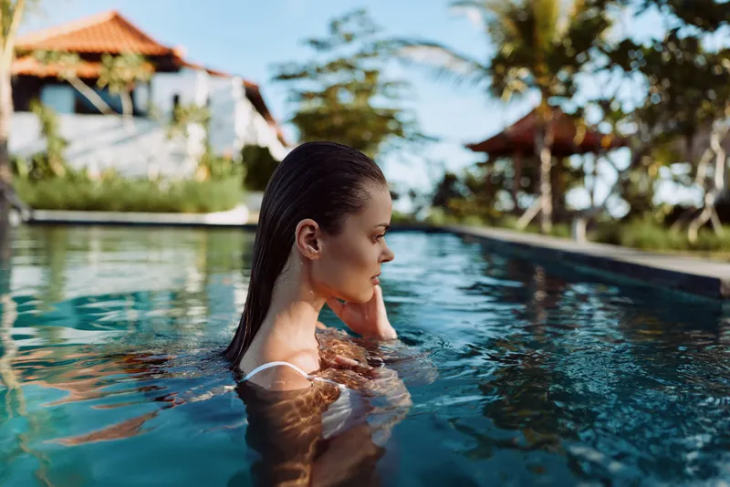 Young woman with wet dark hair relaxes in a sunlit pool, gazing thoughtfully to the side as water mirrors the clear sky, creating a calm, carefree vacation mood and a moment of self care.