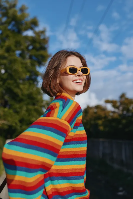 Young woman, a woman wearing a colorful striped sweater and yellow sunglasses, smiling and turning outdoors in a sunny park. Casual outfit, short bob hairstyle, joyful lifestyle portrait.