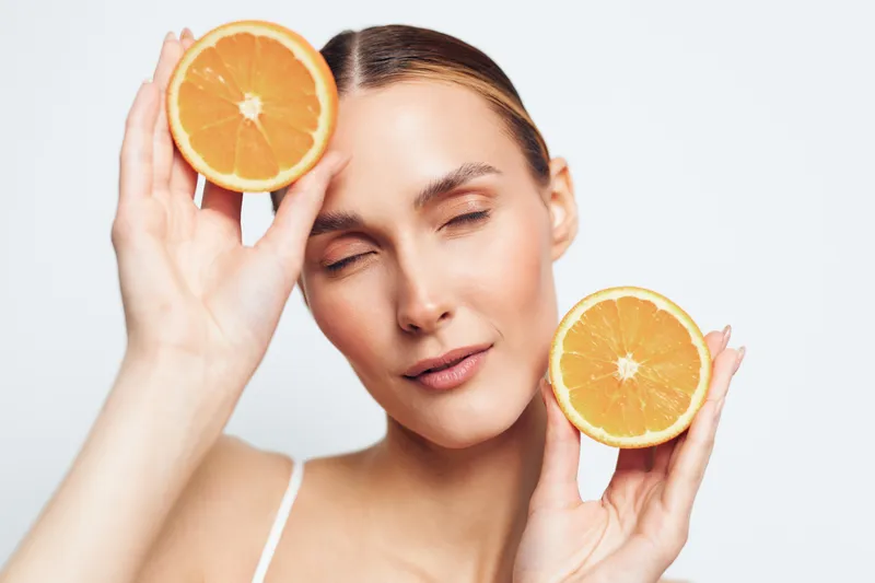 Young woman holds two halves of fresh orange fruit near her face with closed eyes, showing healthy skin and natural beauty on white background, skincare and wellness concept.