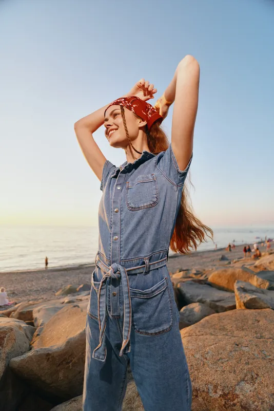 Lifestyle woman in modern boho-western denim street style poses joyfully at sunset on rocky beach with film color tones enhancing warm mood and relaxed vibe.