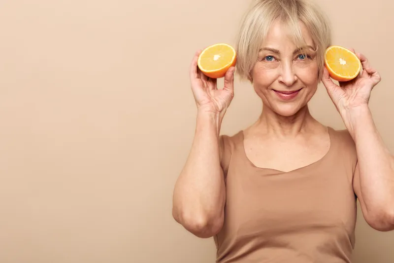 Smiling mature woman holds two orange halves near face on beige background. Concept of healthy lifestyle, vitamin intake, natural nutrition, wellness, and positive mood for seniors.