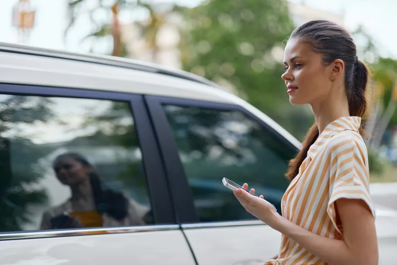 Молодая женщина стоит рядом с автомобилем, увлечённо глядя в смартфон
