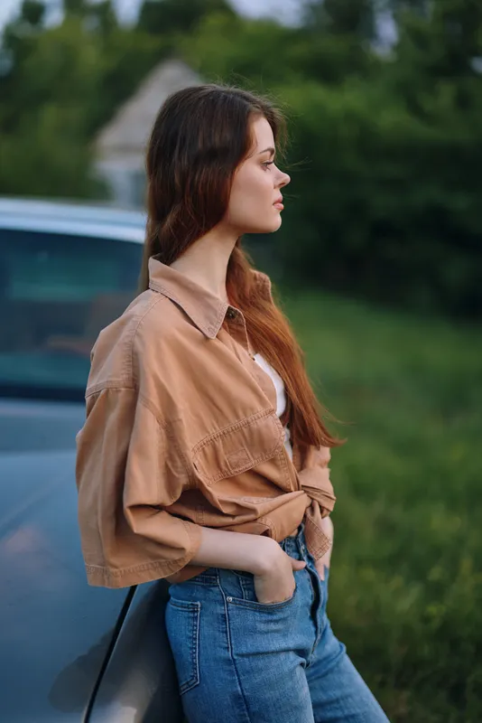 Young woman in trendy casual outfit standing beside a car, showcasing natural beauty and relaxed vibe with a green outdoor background.
