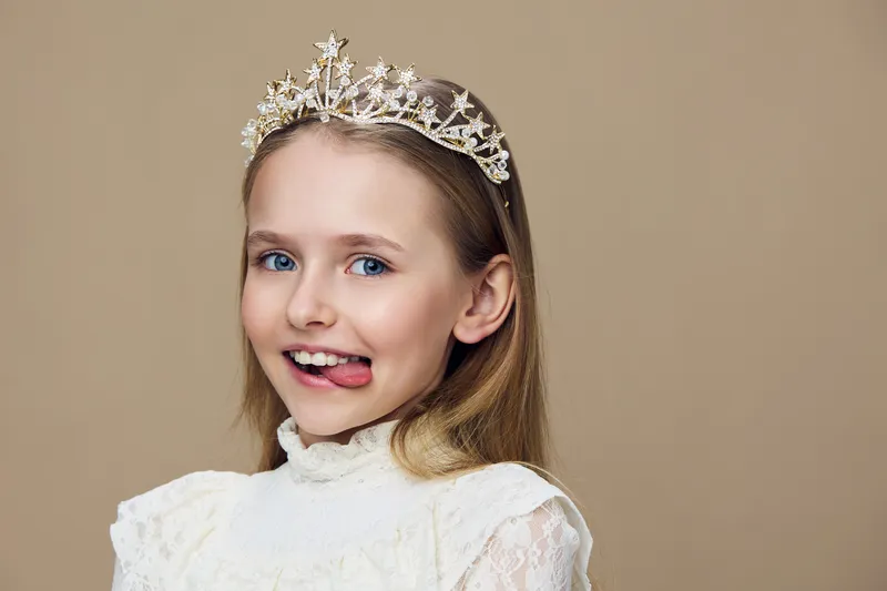 Happy girl with a tiara making a playful face in a light dress, cheerful expression on a soft beige background, perfect for children s themes