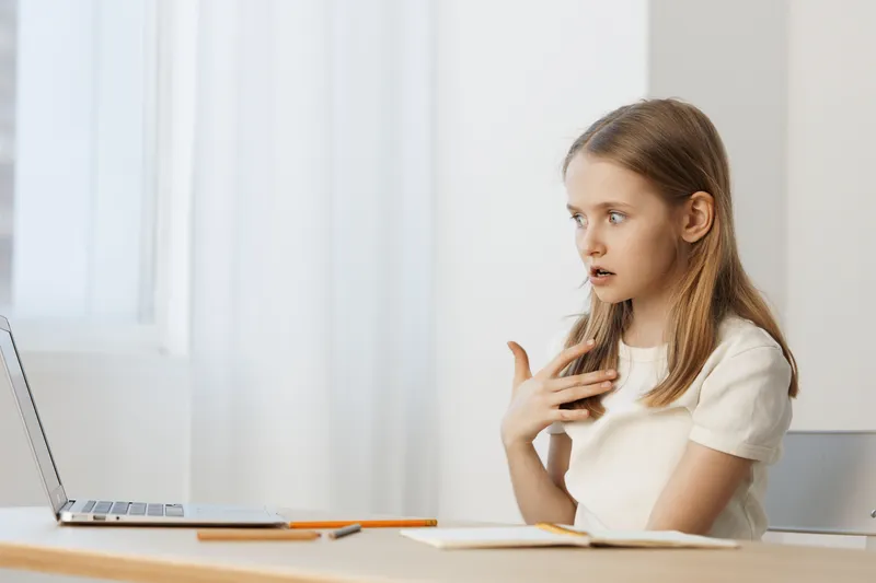 A young girl showing surprise and curiosity while studying at a desk, wearing a white shirt, in a bright and airy room with soft natural light