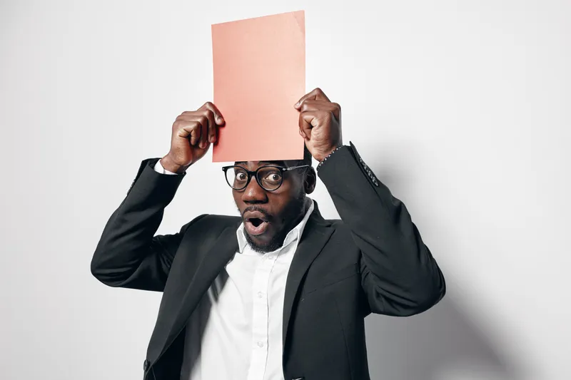 Black man with dark skin wearing glasses and suit holds orange folder above head with surprised expression on white background. Businessman, office, document, emotion, professional.