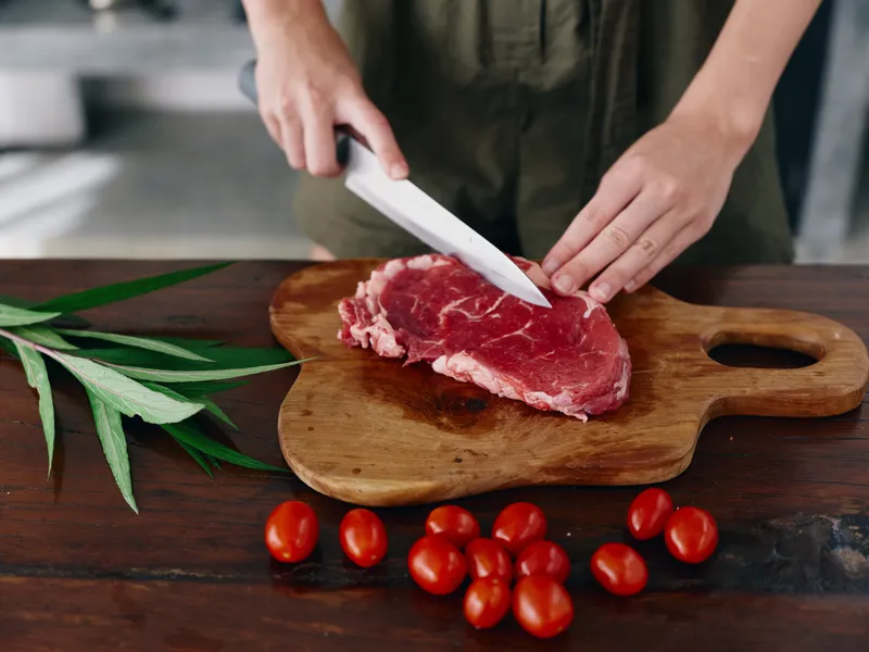 Женщина с ножом в руке нарезает свежее мясо для стейка, предназначенное для запекания на кухне