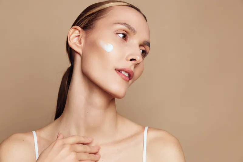 Close up of a young woman applying skin cream on her cheek, soft beige background, natural makeup, gentle glow, skincare routine, healthy beauty concept, serene mood