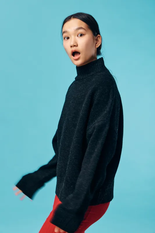 Young woman with surprised expression wearing black sweater and red pants standing against light blue background. Emotion, casual style and portrait in studio with copy space.