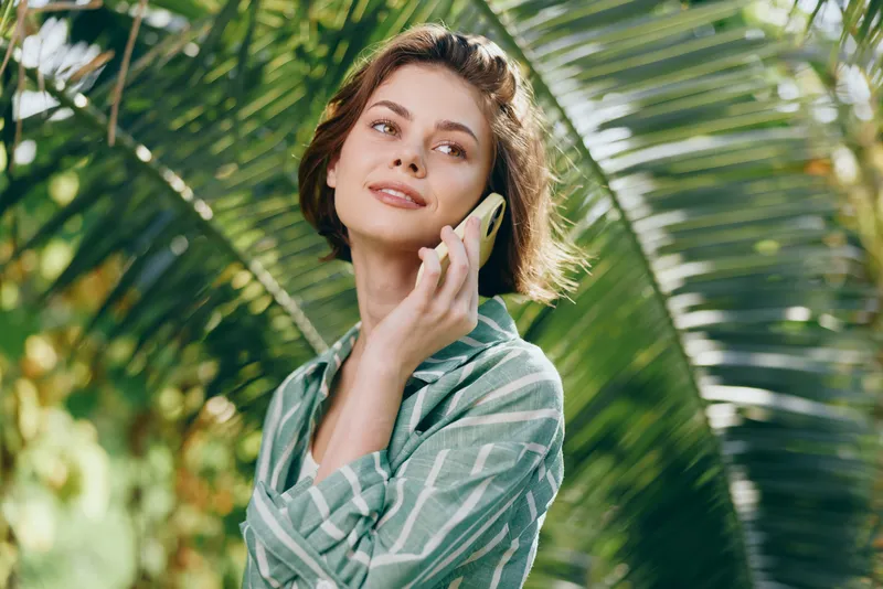 Casual, relaxed woman outdoors, smiling softly while talking on a smartphone among tropical palm leaves and bright greenery.