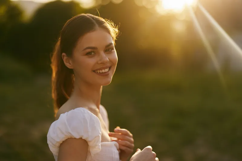 Young woman in white dress smiling during sunset, capturing joy and warmth in a natural background Key themes include happiness, beauty, and nature