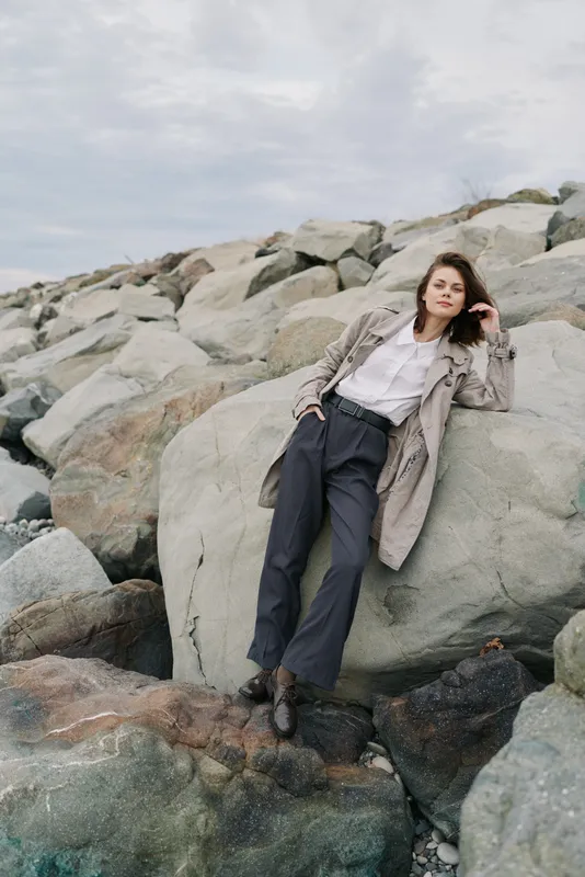 Young woman in casual outfit leans on large rock by rocky shore under cloudy sky. Relaxed pose with hand in hair creates natural lifestyle and outdoor fashion concept.