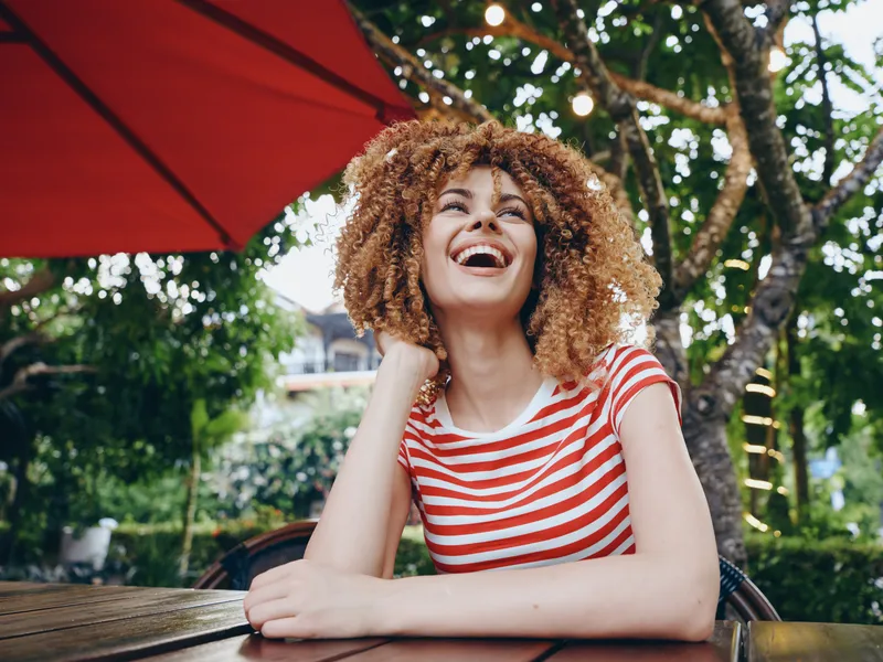 Vibrant portrait of a young woman with curly hair, wearing a striped shirt, laughing at an outdoor cafe. Sunlight, lush greenery, and a bold red umbrella frame a lively moment.
