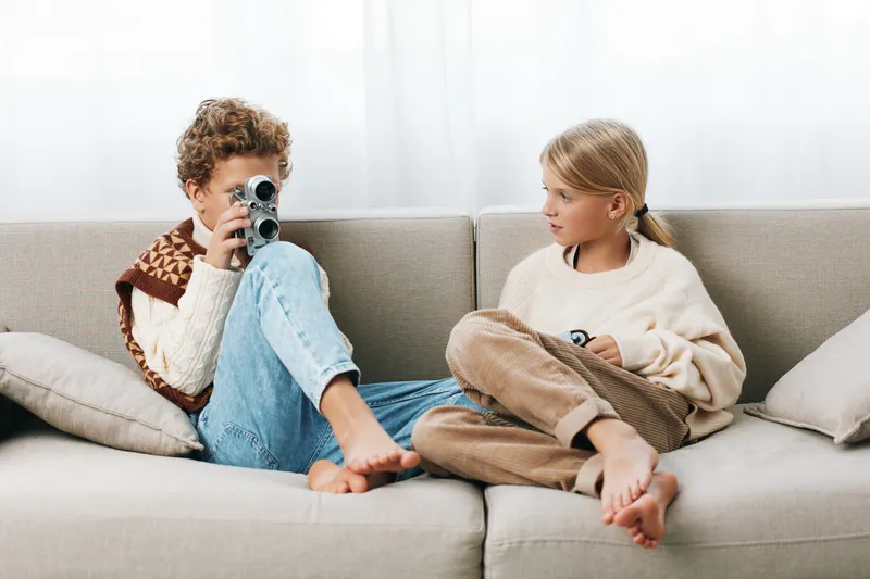 Two children on a soft sofa, a boy with a camera aims and smiles, while a girl sits nearby, creating a warm candid moment of playful curiosity.