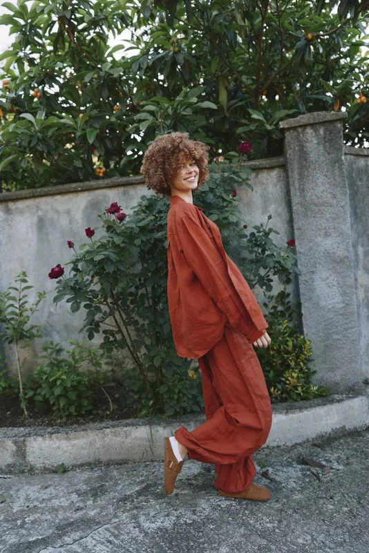Cheerful woman outdoors wearing a rust orange outfit, walking along a garden path with greenery, natural light, candid lifestyle moment, bright and relaxed