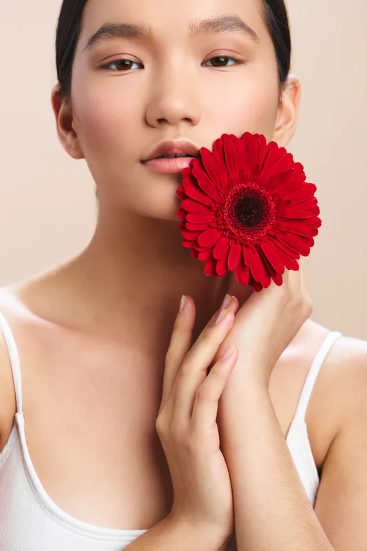 asian woman holds a vibrant red flower near her face, with a soft smile and calm expression, creating a delicate and elegant beauty portrait in warm studio lighting and smooth skin tones.