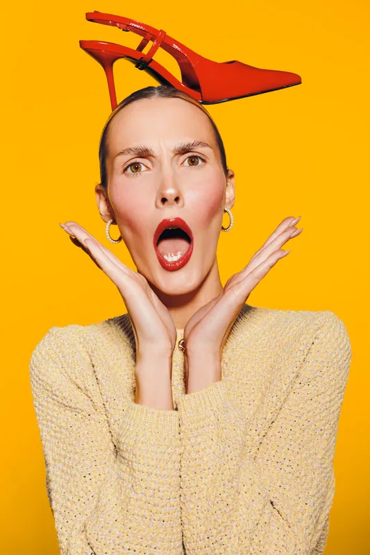 Surprised woman with red lipstick holds red high heel shoe on head against yellow background. Concept of fashion, style, beauty, and creative portrait with bright colors.