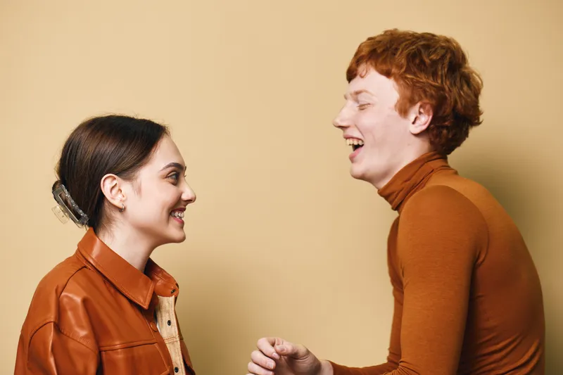 Young man and woman smiling and talking together in casual clothes against beige background. Friendly conversation, positive emotions, interaction and happiness in studio.