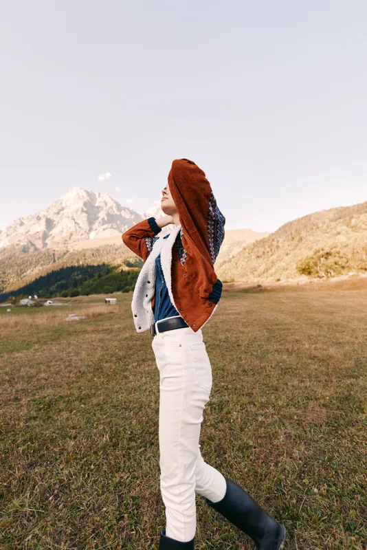 Woman in mountain meadow wearing autumn fashion jacket and boots, portrait of relaxed style outdoors. White pants, knit sweater and scenic landscape for travel and lifestyle.