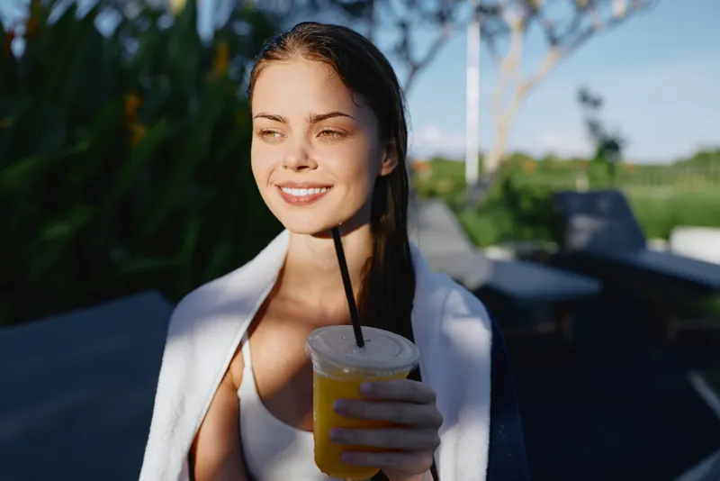 Smiling woman enjoys a refreshing drink outdoors on a sunny day, casual outfit, relaxed mood, natural light, leisure moment captured in a park or resort setting, while holding a cold beverage