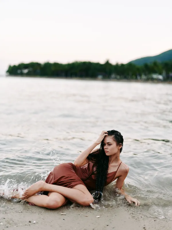 Female figure lounging in shallow sea water at sunset, wearing a warm brown swimsuit, relaxed pose, gentle waves, soft light, reflective mood, beachside calm, introspective moment.