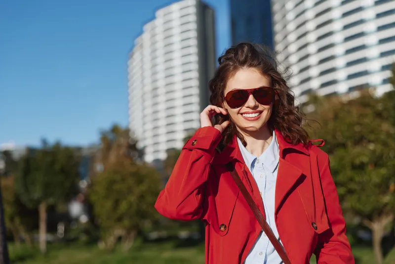 Smiling woman wearing a red coat and sunglasses outdoors in an urban park, bright day. Fashionable portrait with confidence, style, and carefree energy.