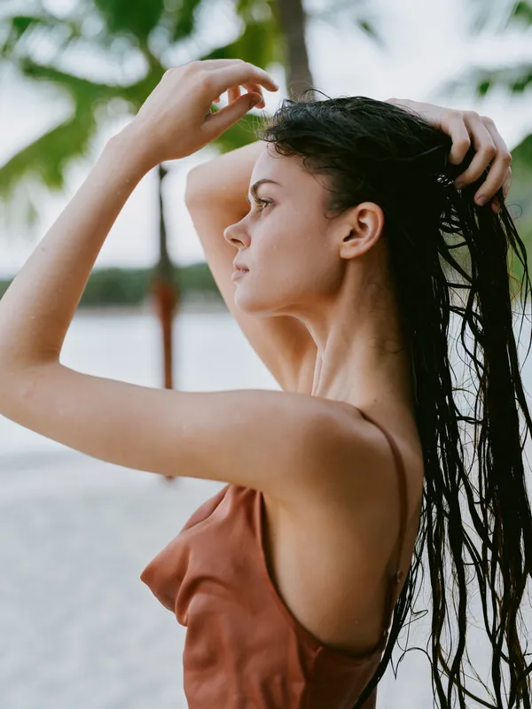 Portrait of a woman on a sunlit beach, rinsing wet hair with a graceful pose. Warm tones, soft light, and water droplets create a calm, natural mood.