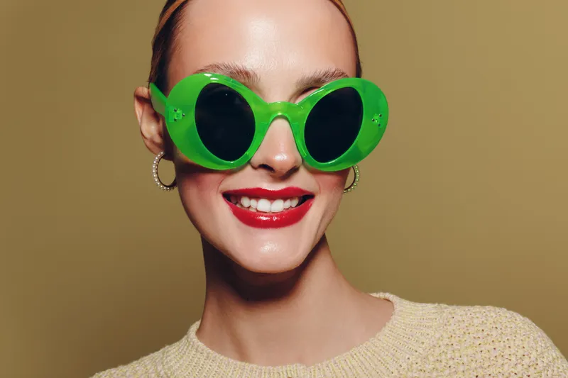 Smiling woman with bright red lipstick wears large green sunglasses and beige sweater. Close-up portrait on neutral background. Concept of fashion, style, and summer accessories.