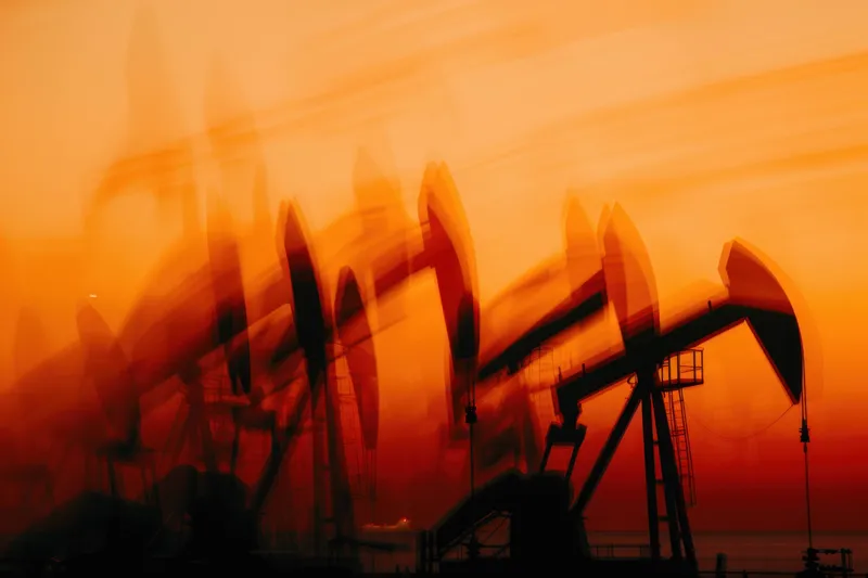 Энергичная сцена с нефтяными вышками в движении, силуэты которых выделяются на фоне поразительного оранжевого и красного заката, олицетворяющая влияние нефтяной промышленности на окружающую среду и…