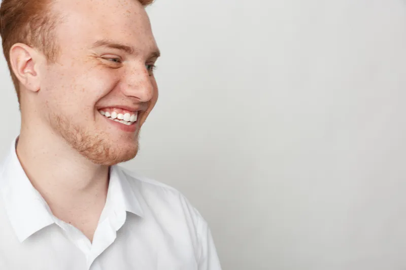 Smiling young man with red hair and beard wearing white shirt looks away on light background. Concept of happiness, casual style, positive emotions, and natural portrait in studio.